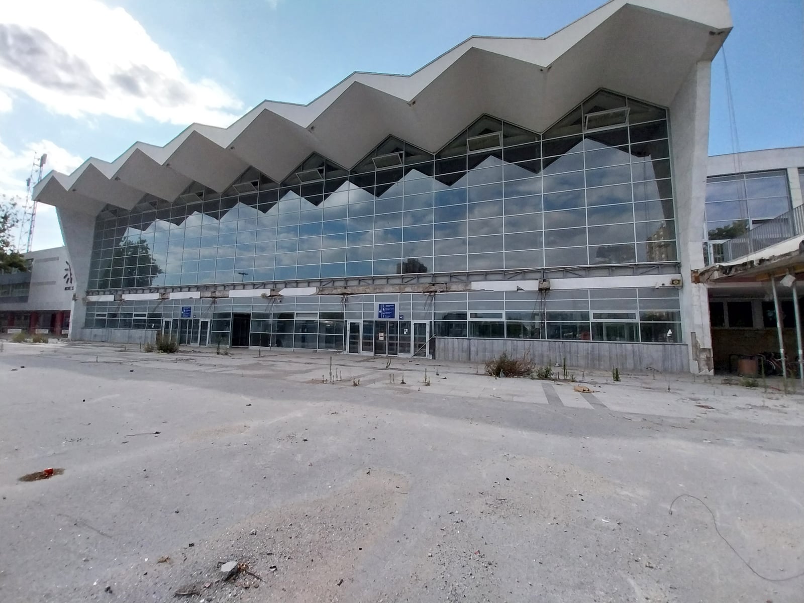The zigzag-roofed glass facade of Novi Sad railway station in July 2026, with no canopy over the entrance and an empty forecourt